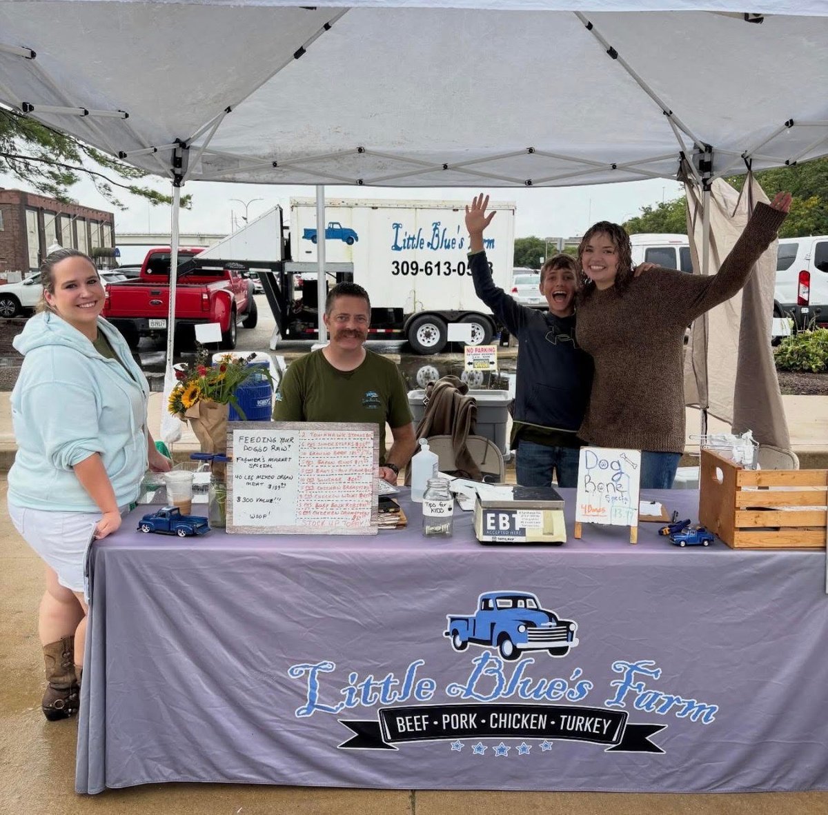 Four people standing behind a market booth under a white tent with a Little Blue's Farm banner displaying meat products