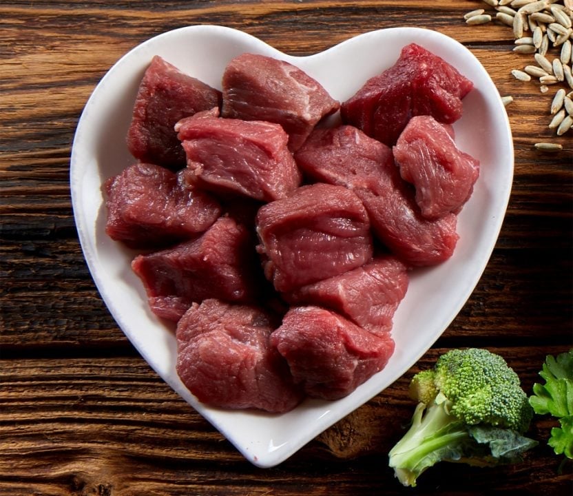 Raw red meat chunks in a white heart-shaped plate on a wooden surface with broccoli and sunflower seeds nearby