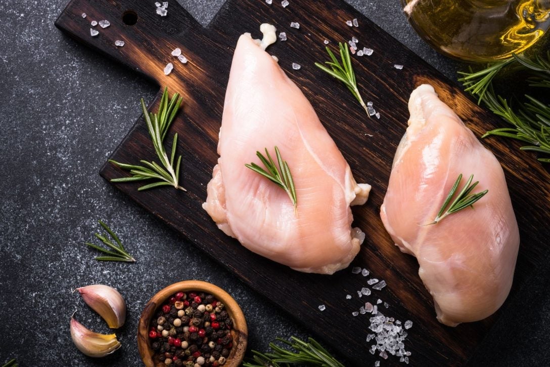 Raw chicken breasts on a dark wooden board with rosemary sprigs, sea salt, peppercorns, and olive oil on a slate background