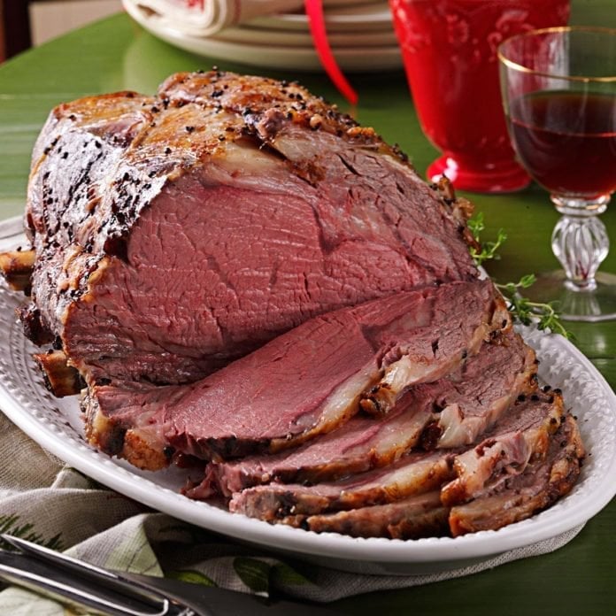 Sliced prime rib roast on white plate with red beverages and place settings on green tablecloth