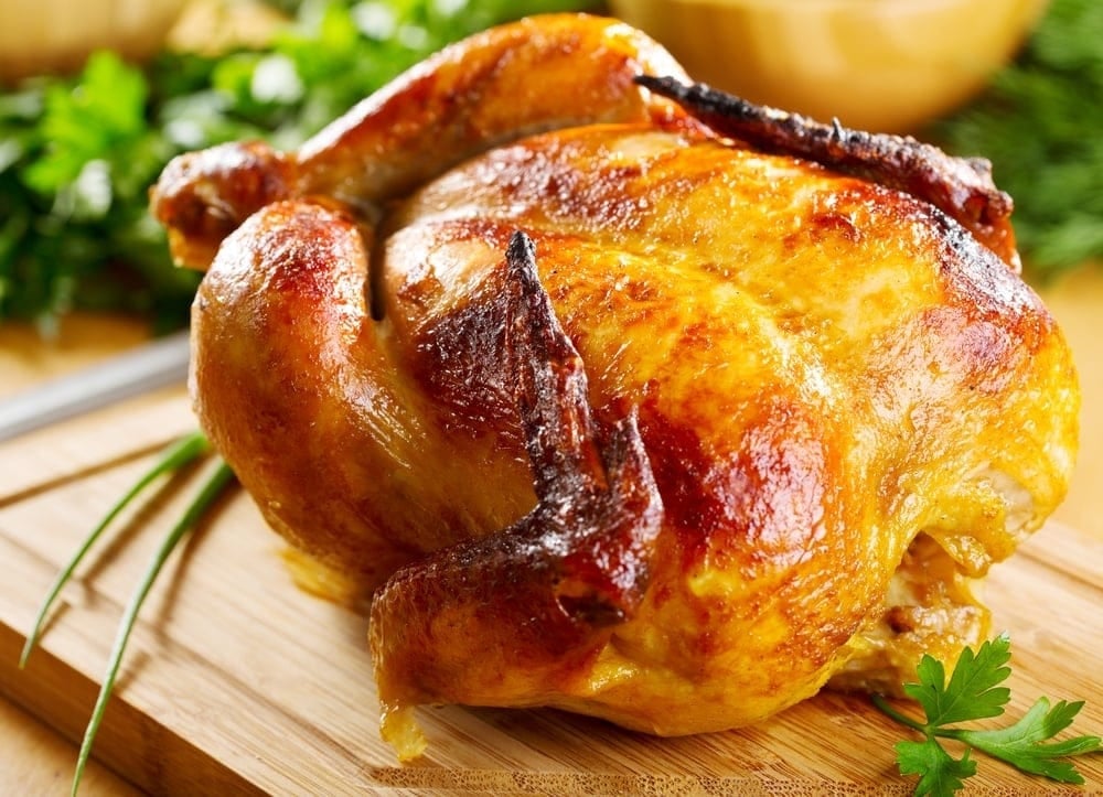 Golden roasted whole chicken on a wooden cutting board with fresh herbs and vegetables in the background